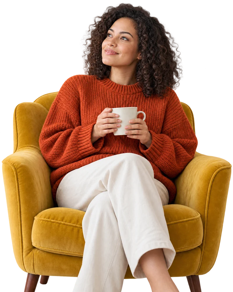 A woman in a rust-colored sweater, holding a mug in a cozy mustard armchair.